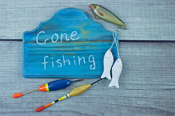 Gone fishing sign written on a pretty wooden plaque. Fishing lures and floats on rustic homemade woody background.Summer fishing flat lay. Top view with copy space