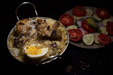Indian spicy dish Biryani with egg, potato and Chicken in a pot along with fresh salads in dark copy space background. Food and object photography.