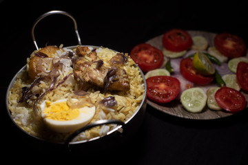 Indian spicy dish Biryani with egg, potato and Chicken in a pot along with fresh salads in dark copy space background. Food and object photography.
