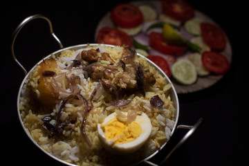 Indian spicy dish Biryani with egg, potato and Chicken in a pot along with fresh salads in dark copy space background. Food and object photography.