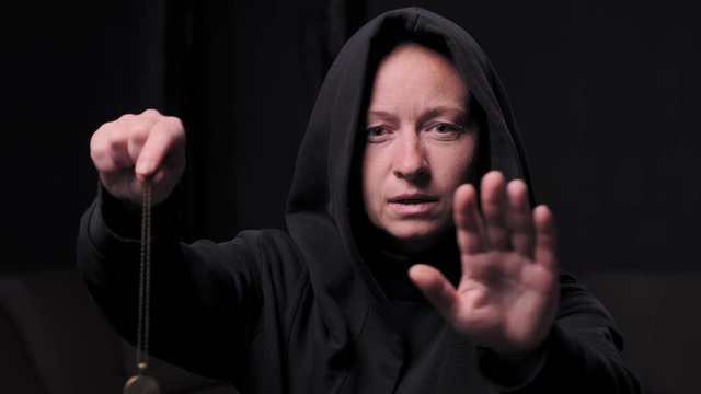 witch woman swings pendant on chain while holding in hand in black room.