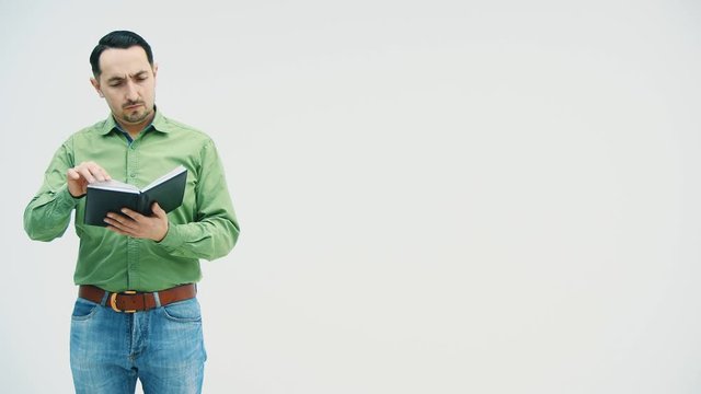 Man standing, reading, checking data in his notebook, then showing opened book to the camera, poiting with finger at what is writen there. Funny face expression.
