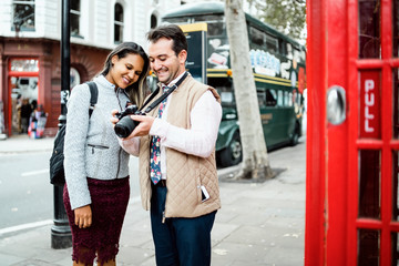 Fototapeta premium Happy travelling couple reviewing photos in London, UK. Double decker bus and red telephone booth in a frame.