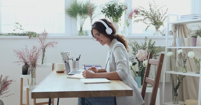 Female florist designer wears headphone elearning online watching video training course on laptop at work surrounded with flowers. Wedding planner video conferencing client in modern floristry studio.