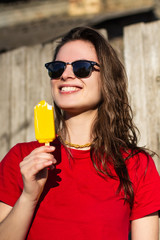 Teenage girl with yellow ice cream outdoors. Young brunette woman in sunglasses and red t shirt against wooden wall fence eating delicious cream. Concept of dessert, emotions, joy, teenage lifestyle.