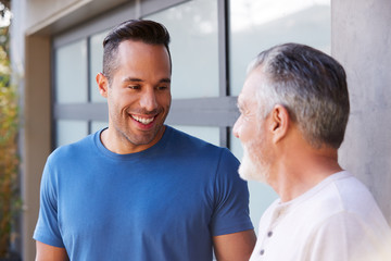 Senior Hispanic Man Talking And Laughing With Adult Son In Garden At Home