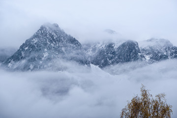 Overcast autumn alpine view with mountain silhoette fragments through fog and clouds. Climate, environment and weather concept sky background.