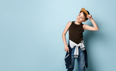 Active kid dressed in sporty style stands looking aside and touching cap on his head, creating...