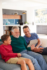 Multi-Generation Male Hispanic Family Sitting On Sofa At Home Watching Movie On Digital Tablet