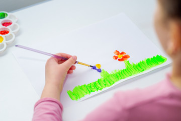 Hand of child drawing flowers on green grass with brush by watercolors on white paper.