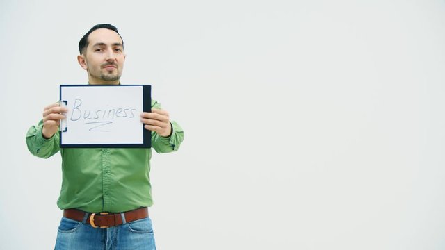 Funny young man standing holding the paper holder with word "bisiness" writen on it, showing it at the camera, pointing at it, playing, moving comically.