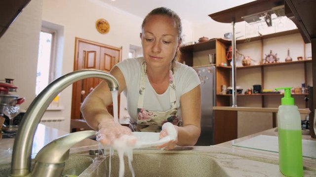 woman washes dishes in kitchen. Housewife rinses plate under tap water in sink.