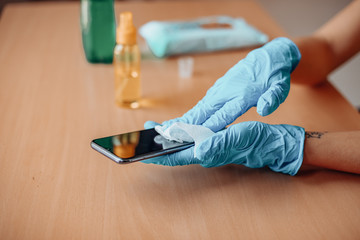 Woman's hand in blue gloves sanitizing cleaning smartphone mobile phone with alcohol on wood table surface with wet wipes