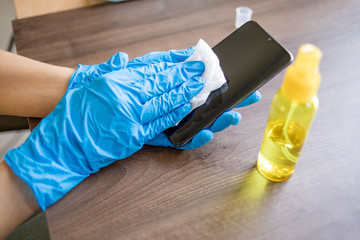 Woman's hand in blue gloves sanitizing cleaning smartphone mobile phone with alcohol on wood table surface with wet wipes