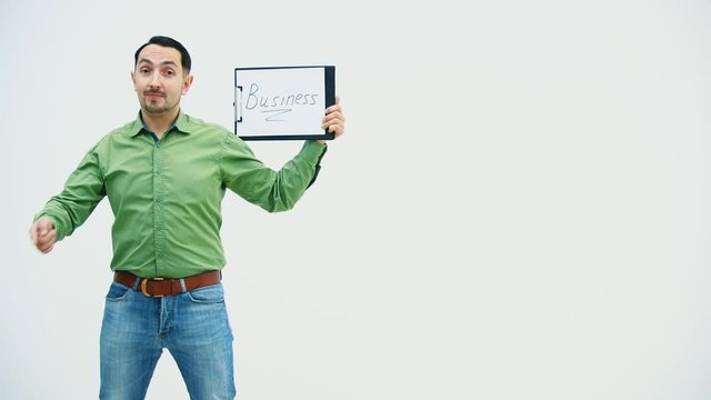 Funny young man standing holding the paper holder with word "bisiness" writen on it, showing it at the camera, pointing at it, behaving comically.