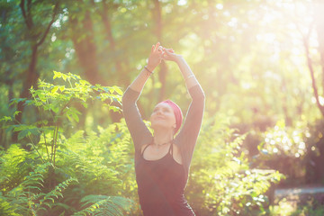 woman stands in the forest at sunset. haze of sunlight. hands raised connected. looking up.