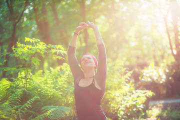 woman stands in yoga pose in the forest at sunset. haze of sunlight. hands raised looking up.