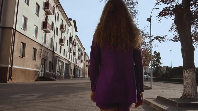 Business lady concept. Curly long haired girl in a suit walks along city street, rear view on the back. Vogue female goes alone. Fashionable stylish woman moves in slow motion. Trendy lady is outdoors