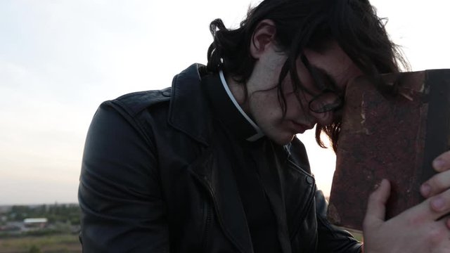 Handsome young priest portrait outside at sunset. He is wearig eye glasses and roman collar shirt and a leather jacket.