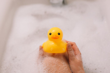 Girl is holding yellow rubber duck in the bath with bubble foam