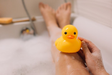 Girl is holding on her legs yellow rubber duck in the bath with bubble foam