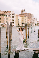 Italy wedding in Venice. The bride are standing on a wooden pier for boats and gondolas, near the Striped green and white mooring poles, against backdrop of facades of Grand Canal buildings.