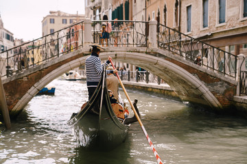 Italy wedding in Venice. A gondolier rolls a bride and groom in a classic wooden gondola along a narrow Venetian canal. Gondola swims under an arched bridge, rear view.