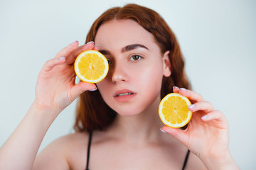 portrait of redheaded young woman standing on isolated white background holding half of orange in hand closing her eye with another half , beauty portrait concept