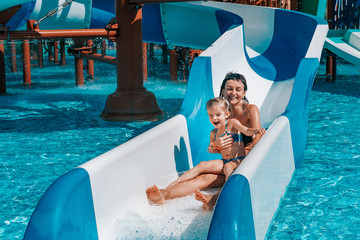 Little girl in swimsuit goes down the blue slides to the pool, Mom and daughter play and swim in...