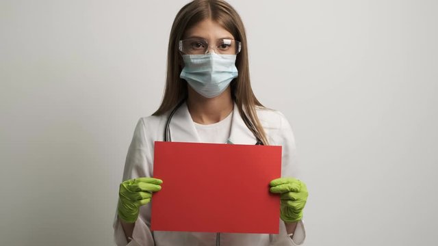 A female doctor in a mask of Caucasian appearance holds a red A4 sheet in her hands