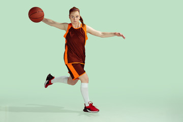 Young caucasian female basketball player in action, motion during gameplay isolated on mint colored background. Concept of sport, movement, energy and dynamic, healthy lifestyle. Training, practicing.