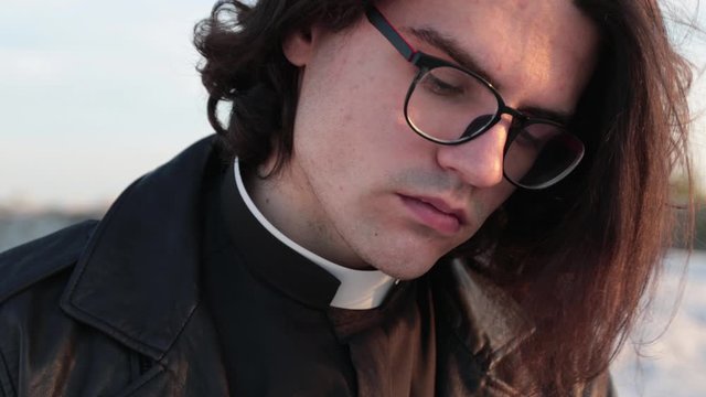 Handsome young priest portrait outside at sunset. He is wearig eye glasses and roman collar shirt and a leather jacket.