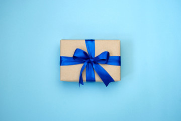 Gift box with blue ribbon bow wrapped in kraf paper on blue background. Present for Father's Day, Birthday or international Men's Day. Festive decor background. Top view layout with copy space. 