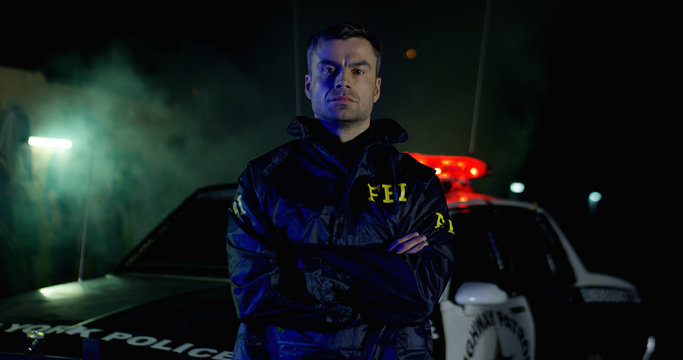 Portrait of police officer with serious faces looking at camera. FBI agent work at the scene at night, police car with lights on background.