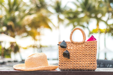 Weaving bag and hat with sunglasses and sunscreen lotion next to a swimming pool, summer travel concept, coconut tree at a beach background