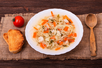 Fresh chicken soup with noodles and various vegetables in a white plate served with slices of toast on a textured burlap, angle view