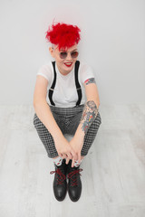 Portrait of a cool fashionable modern young girl. A short haircut with shaved temple. Dyed bright red hair. Red lipstick. Studio photo on white background. Suspenders and checkered pants.