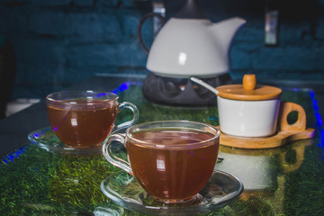 Tea-set. In front are two cups of tea. Behind the sugar bowl and kettle. On a glass table