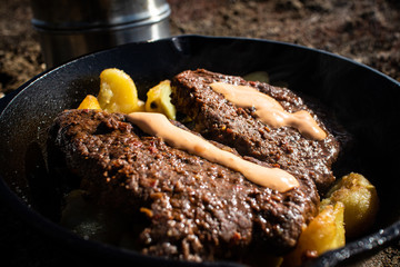 Cooking steaks and potatoes with cast iron frying pan next to sand pit at spring time.