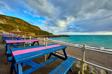 Image of Bouley Bay with harbour in the background, the beach and cafe benches in the foreground. Jersey Channel Islands
