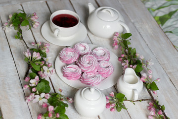 breakfast romantic decoration with pink marshmallows on plate with teapot, cup of hibiscus tea, milk and sugar jugs, blooming apple tree branchs on white wooden table