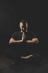Fototapeta na wymiar Young Caucasian man sitting with hands in namaste gesture with black clothes and black background.