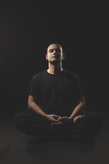 Young Caucasian man sitting in meditation with black clothes and black background.