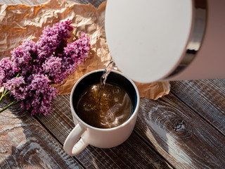 Boiling water from an electric kettle pours into a Cup on a wooden background with a bouquet of lilacs.
