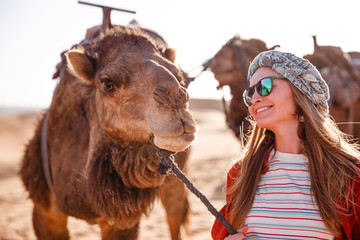 Beautiful young slim woman in a turban and sunglasses communicates with a camel at dawn in the Sahara desert. Morocco.
