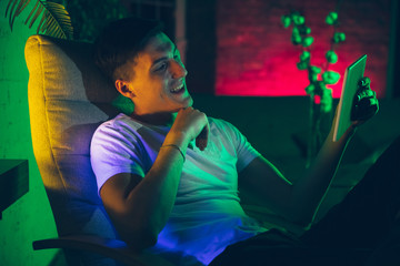 Laughting. Cinematic portrait of stylish man in neon lighted interior. Toned like cinema effects, bright neoned colors. Caucasian model using gadgets, devices in colorful lights indoors. Youth culture