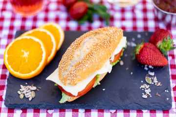Wooden table with white and red rustic napkin. A seed sandwich stuffed with eggs, cheese, tomato and lettuce. Some fruit and a glass of wine