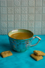 Closeup view of a cup full with black coffee and small cookies laying on turquoise surface against white ceramic tiles in kitchen
