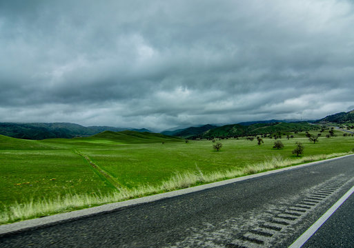 Field with green grass, asphalt road with a white line, mountains on the horizon, cloudy blue sky. The usual landscape. Beauty of nature. Beautiful horizontal photo in good quality.
