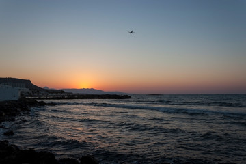 Sunset over the sea in Kokkini Hani, Crete, Greece.  The sun disappears behind the mountain. A plane is flying in the sky. Scenic seaside landscape in the evening.